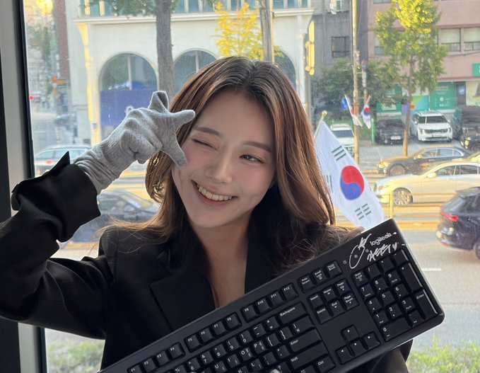 A smiling young woman winks at the camera, holding a black Logitech keyboard signed with 'IVE' and 'Liz' alongside a small South Korean flag, wearing a silver glove on one hand.