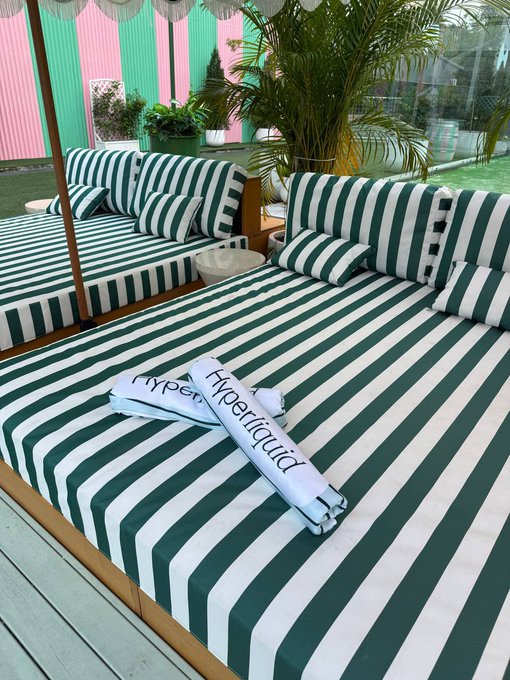 Outdoor lounge area with green and white striped daybeds and umbrellas, featuring two Hyperliquid branded towels placed on the cushions.