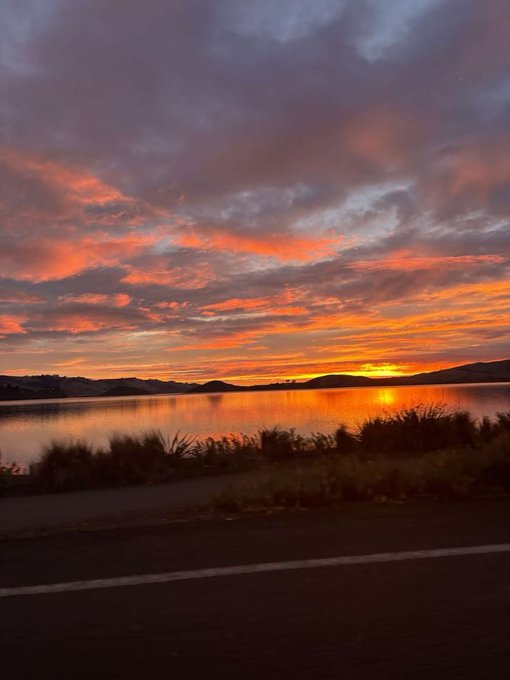 A vibrant orange and red sunset reflects across a calm lake with silhouetted mountains in the distance, viewed from beside a road.