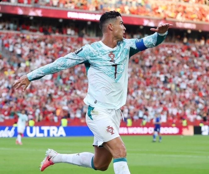 Soccer player Cristiano Ronaldo, wearing a white and teal Portugal jersey with the number 7, celebrates a goal with his arms outstretched in a crowded stadium.