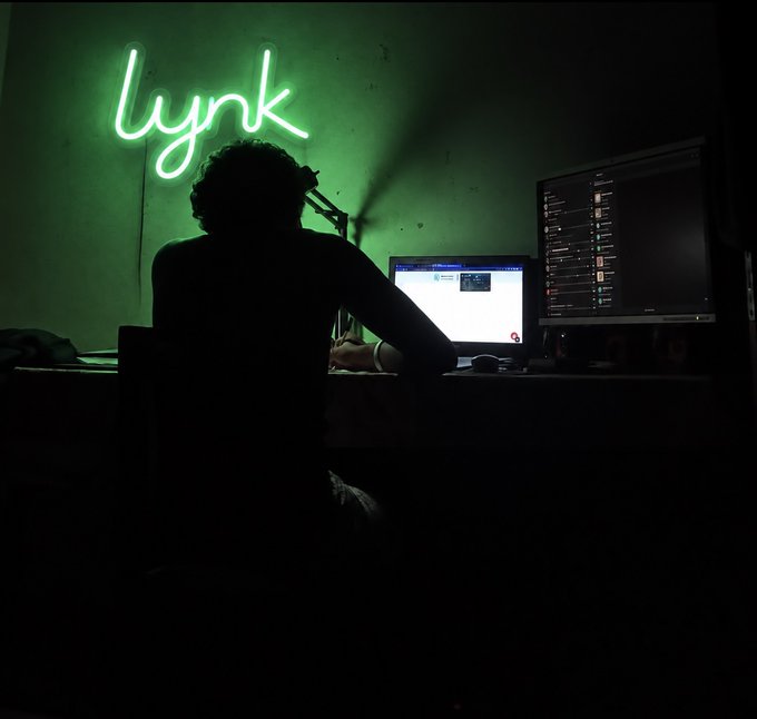 A silhouetted figure sits at a desk working on a laptop and monitor, illuminated by a glowing green neon "lynk" sign on the wall. The dark, focused setup reflects a late-night "work in" session.