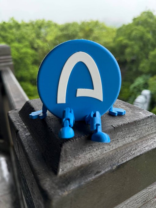 A blue, egg-shaped character figurine with a white letter 'P' on its face and small legs sits on a dark wooden railing with green trees blurred in the background.