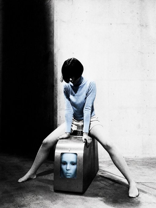 A black-and-white photo, accented with a blue tint on the sweater, depicts a person sitting astride a metal box that displays a glowing blue face on a screen.