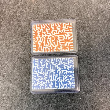 Two cassette tapes or similar items with abstract line-art patterns, one orange and one blue, resting on a gray carpet.
