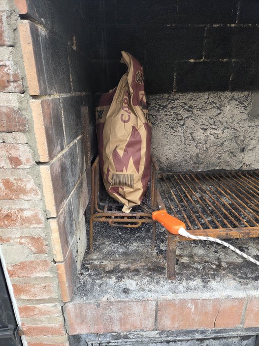 A large crumpled brown paper bag with purple graphics sits on a rusty metal grill grate inside a brick barbecue pit, with a white and orange tool resting on the grate.