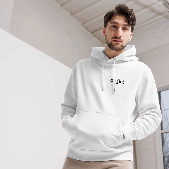 A young man modeling a white hoodie with the text 'oajkt' printed on the chest in a bright, minimalist indoor setting.