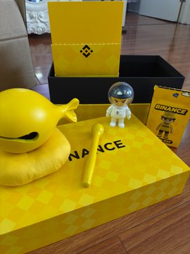 The image features a yellow box with a black lid, which appears to be a toy or game set for children. The box contains various toys such as action figures, a toy boat, and other items. There are also two small figurines of people in the scene, one on top of the box and another on the floor next to it. The post includes text that reads "出了社会后，只有币安儿童节 @binancezh 早上收到了 币安儿童节玩具", which seems to be a reference to the toy set in the image.