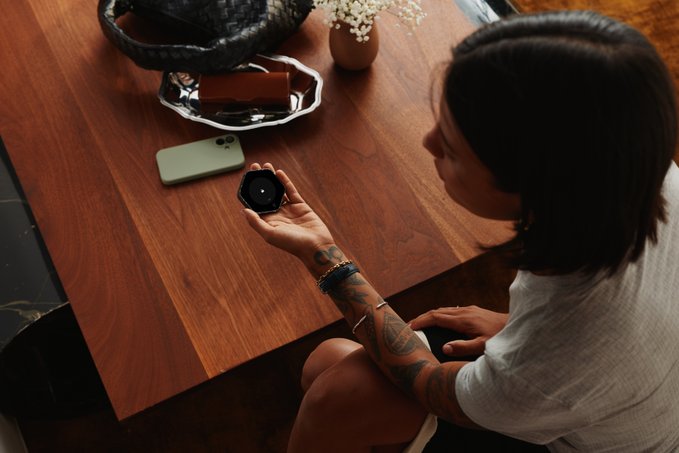 A woman with dark hair and a tattooed arm holds a black smart device above a wooden table, next to a green smartphone and a black woven bag.