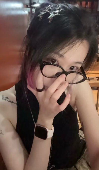 A young woman with dark hair featuring pink highlights poses indoors, holding black-rimmed glasses to her face, wearing a black top, a star-shaped hair accessory, and a smartwatch.