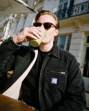 A photograph of a person wearing sunglasses and a black jacket drinking from a paper cup while seated at an outdoor café.