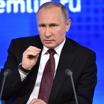 Russian President Vladimir Putin wearing a dark suit with a burgundy tie, gesturing with his hand at a podium with microphones, with Cyrillic text visible in the background.