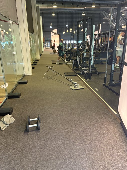 A spacious, modern gym with industrial-style lighting and various weightlifting equipment, including a cable machine, weight plates, and a barbell on the floor. A man is seen walking through the open workout area, and the post mentions "Gym time" alongside an @unvault Phase 1 giveaway.