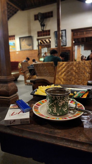 The image shows a coffee cup sitting on top of a table, with a book next to it. The scene takes place in an indoor setting where people are seated at tables or benches, possibly enjoying their time together. There are also chairs placed around the room. A person is visible in the background, likely engaged in conversation or other activities.