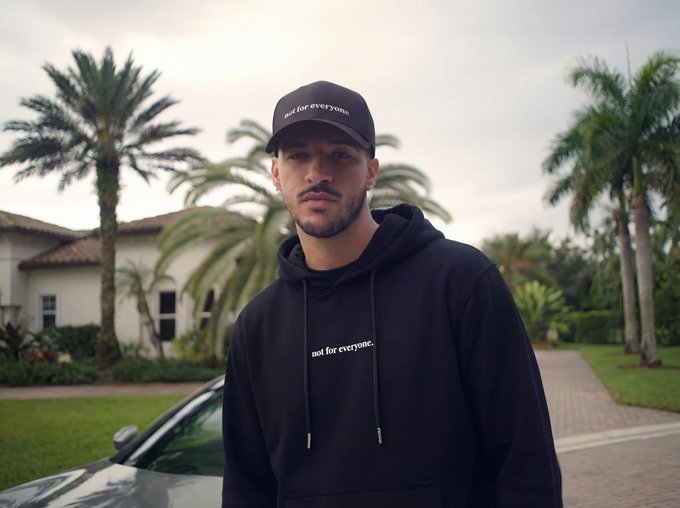 A man wearing a black cap and black hoodie with 'not for everyone.' text, standing in front of palm trees and a house with a car partially visible.