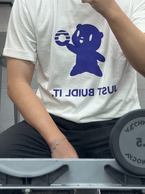 A mirror selfie showing a person wearing a white t-shirt featuring a blue cartoon bear holding a coin, with text reading 'JUST BUND IT.' (shown reversed) and a Technogym dumbbell in the foreground.