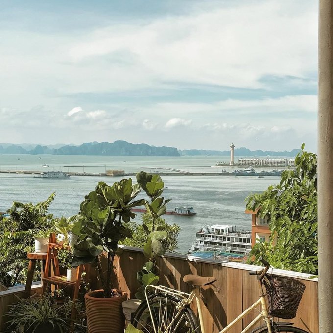 A scenic view from a balcony with potted plants and a bicycle, looking out over a harbor with boats, a lighthouse, and limestone karst islands in the distance.