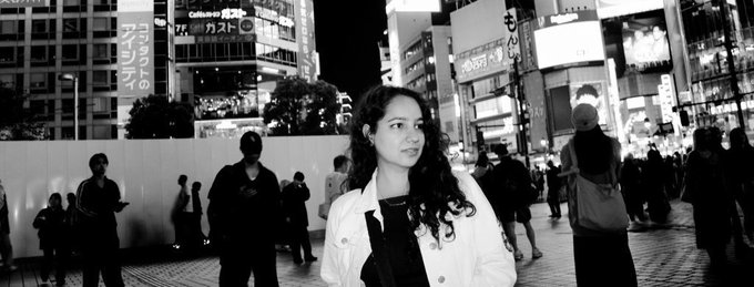 A black-and-white street photograph of a young woman in a white jacket standing in a busy Japanese urban area at night with neon signs.