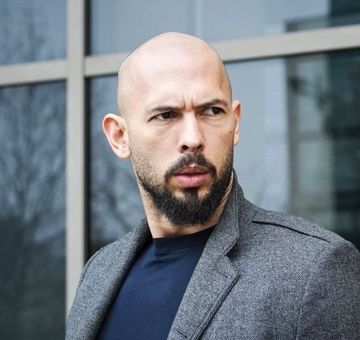 A bald man with a beard wearing a gray blazer over a dark blue shirt looks off to the side in front of a building with large windows.