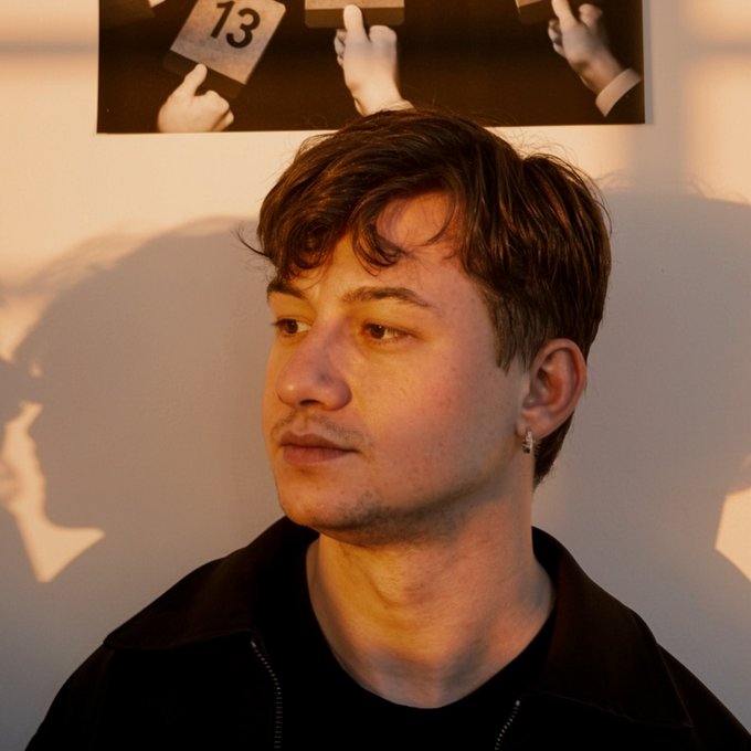 A portrait of a young man with dark, wavy hair bathed in warm, golden sunlight, with a partially visible poster featuring the number '13' and hands in the background.