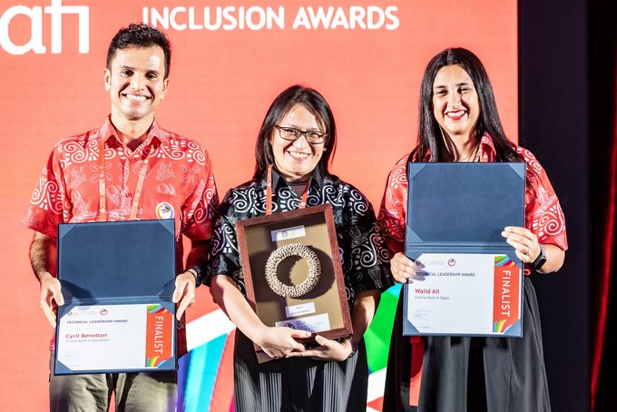 Three people stand on a stage smiling and holding awards in front of a backdrop reading 'afh INCLUSION AWARDS'. Two finalists hold blue folders; the man on the left is named 'Cyril Benediction' and the woman on the right is 'Wahid Ali'. The woman in the center holds a decorative spiral trophy.