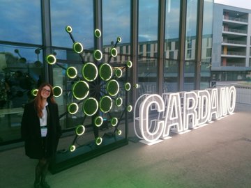 A young woman poses in front of an illuminated 'CARDANO' sign and a green circular sculpture, likely at a Cardano-related event.