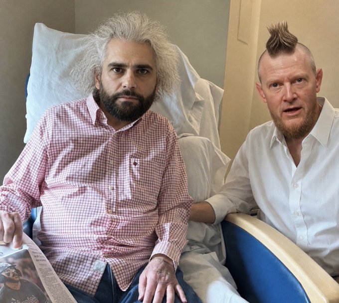 Two men sit in a hospital room, one in a plaid shirt in a hospital bed and another in a white shirt with a mohawk beside him, with what appears to be a manga visible in the foreground.
