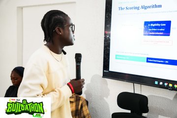 A person with braided hair and glasses speaks into a microphone in front of a screen titled 'The Scoring Algorithm,' with a 'Buildathon JOB' logo in the bottom left corner.