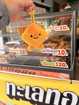 A hand holds up a small, fuzzy yellow plush keychain with a smiling face above a food stall displaying fried chicken with Thai pricing labels (10฿ and 120฿).