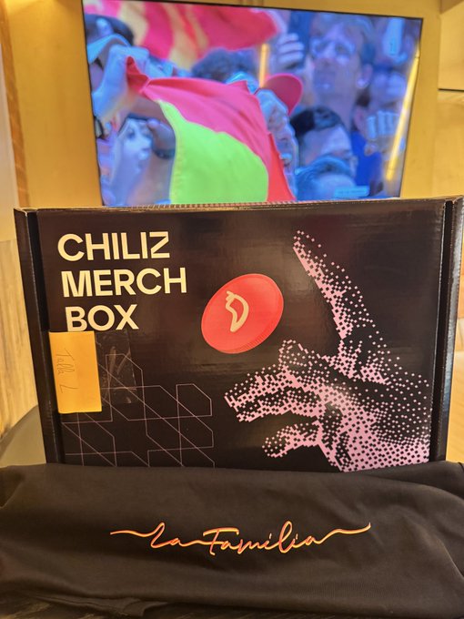 A photo showing a 'CHILIZ MERCH BOX' with the Chiliz logo placed in front of a TV displaying a sports crowd scene, with a black 'La Familia' cloth below.