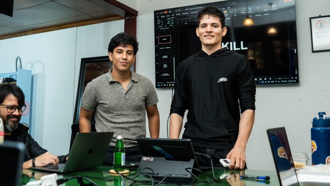 Three men are gathered around a table with laptops in a workspace; two stand smiling at the camera while a third sits to the left, with a monitor displaying 'KILL' visible behind them.