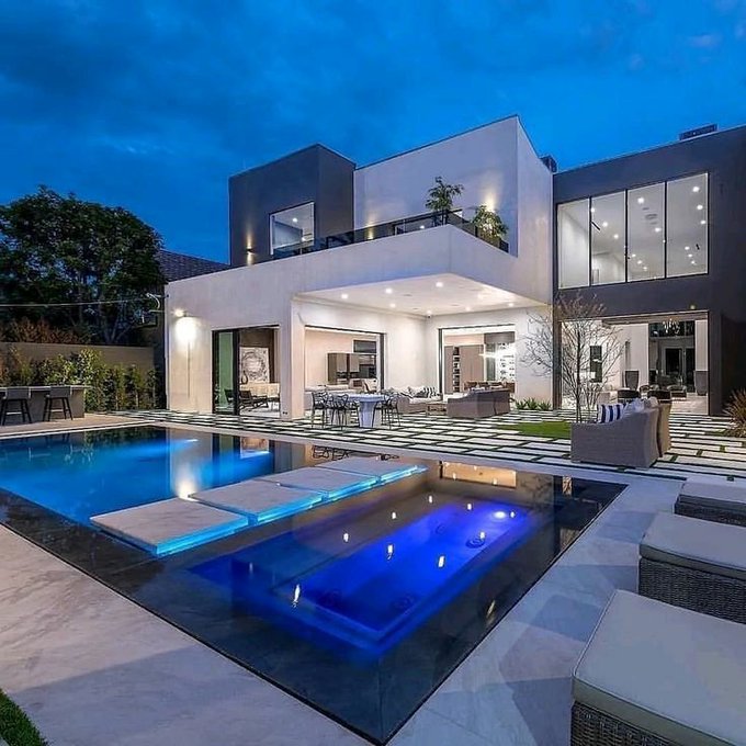 A luxurious modern mansion photographed at dusk featuring a contemporary white and dark gray facade, illuminated outdoor patio with seating areas, and a stunning swimming pool with blue-lit water and floating platforms.