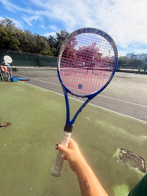 A hand holds up a HEAD brand tennis racket on an outdoor court, ready for a workout—embodying the "health is wealth" message through active play under a sunny sky. The racket's pink strings and blue frame stand out as a symbol of fitness investment.