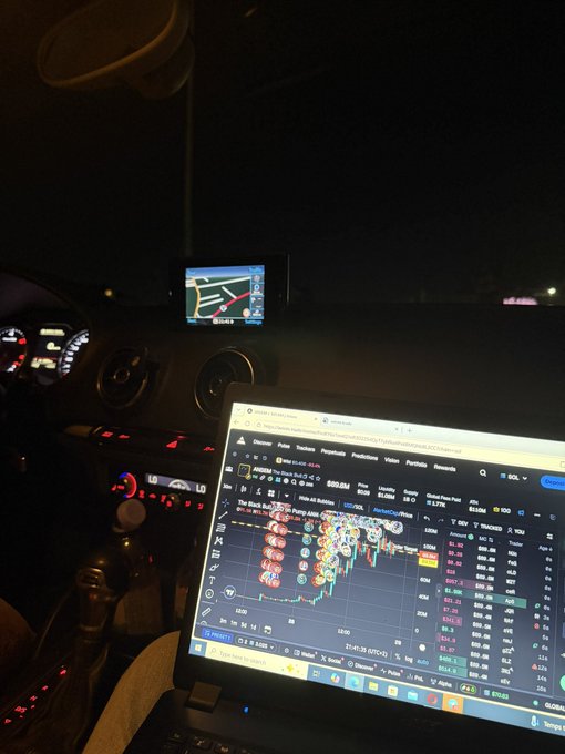 A laptop inside a car displays a Solana SOL/USD trading chart ($229.54) alongside a GPS navigation screen, illustrating a late-night "onboarding" session as the person works outside the vehicle to set up a crypto wallet for a friend in what appears to be a parking lot.