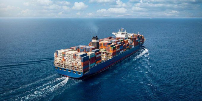An aerial view shows a massive blue cargo ship heavily loaded with colorful shipping containers sailing across the deep blue ocean under a partly cloudy sky.