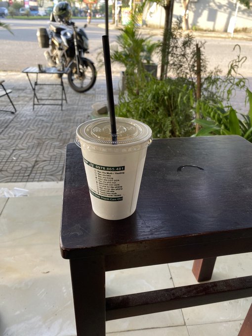 A plastic cup of iced coffee with a black straw sitting on a dark wooden stool on a sidewalk, with a motorcycle and street visible in the background.
