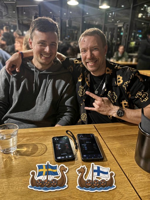 Two smiling men pose together at a wooden table in a restaurant or bar setting; one wears a grey hoodie, while the other wears a black shirt with gold Bitcoin patterns and makes a hand gesture. Two smartphones, a glass of water, and stickers of Swedish and Finnish Viking ships are visible on the table.