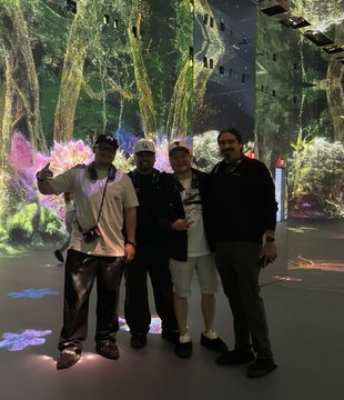 Four men posing for a group photo in a modern, dimly lit art space featuring immersive, glowing digital projections of a forest and flowers on the walls and floor.