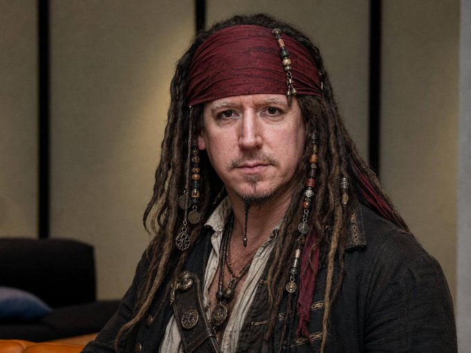 A man with long dreadlocks, a red bandana, and beaded accessories wearing a pirate-style outfit, resembling Captain Jack Sparrow.