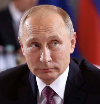 A close-up portrait of a man in a suit with a purple tie, with a Russian flag visible in the background.