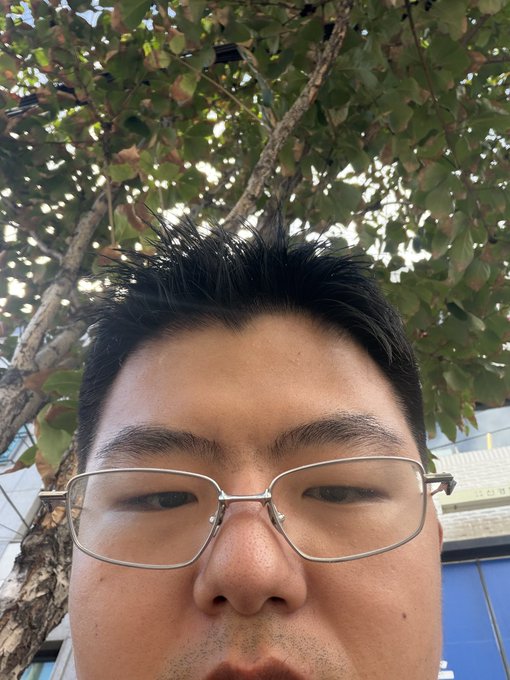 A close-up selfie of a man wearing rectangular glasses, taken beneath leafy tree branches, visually captures the intense, scrutinizing focus of a trader monitoring rapid market movements. The composition mirrors the post's sentiment of being overwhelmed yet hyper-attentive, as his narrowed, concentrated gaze seems locked on an unseen screen reflecting fast-paced trading action.