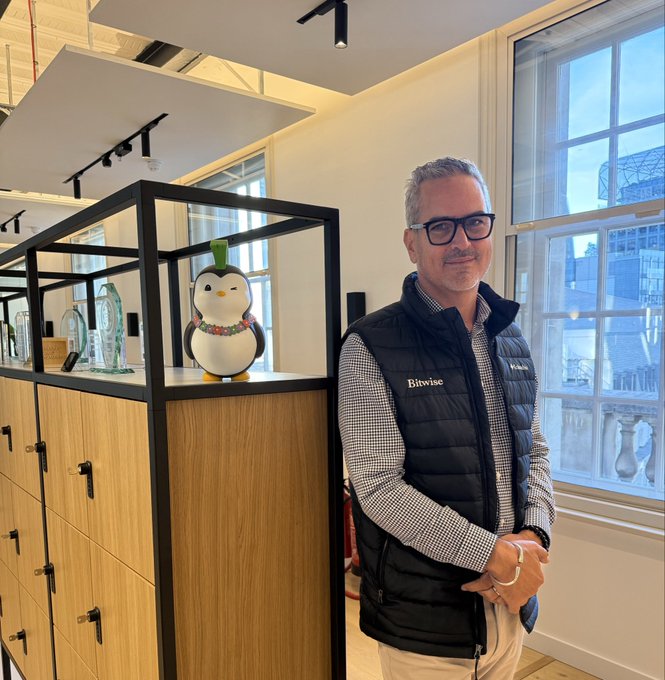 A man wearing a "Bitwise" branded vest stands beside a display case featuring a collectible penguin figure with a green tuft of hair and a colorful lei, reflecting the post's celebration of personalized NFT Penguin characters.