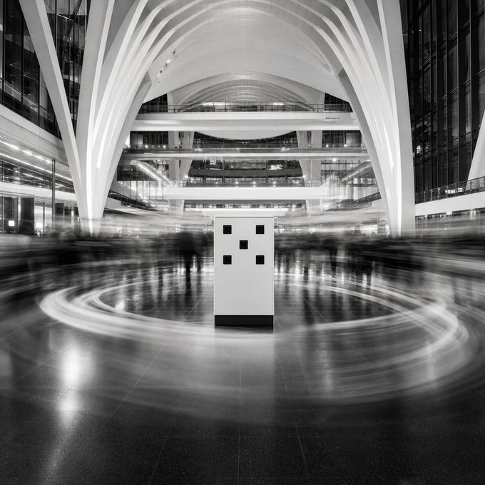 A black and white long-exposure photograph of a modern building's grand interior with sweeping architectural arches and blurred figures circling around a central pillar with square cutouts.