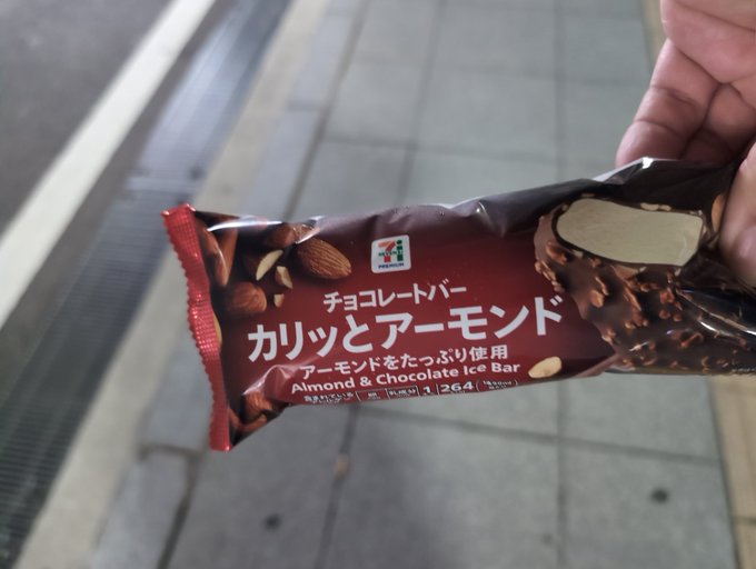 A hand holds a 7-Eleven Almond & Chocolate Ice Bar (チョコレートバー カリッとアーモンド) in front of a tiled sidewalk.