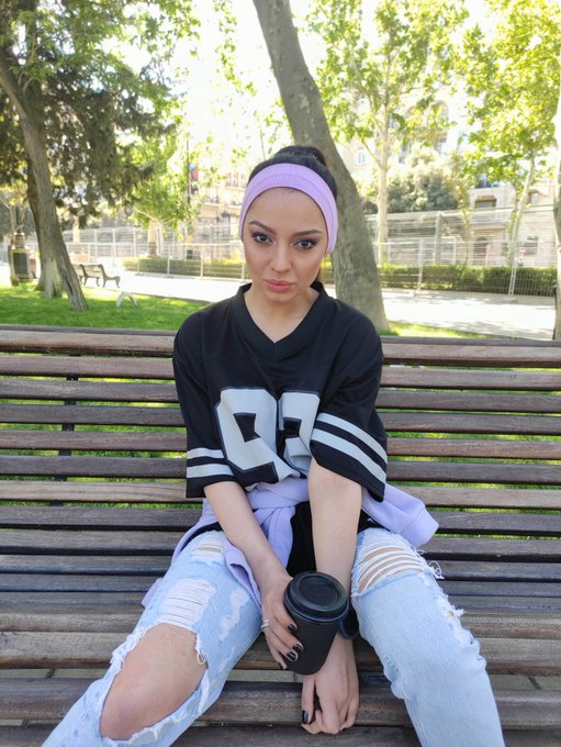 A young woman wearing a lavender headband, black jersey with the number '87', and ripped jeans sits on a wooden park bench holding a coffee cup.