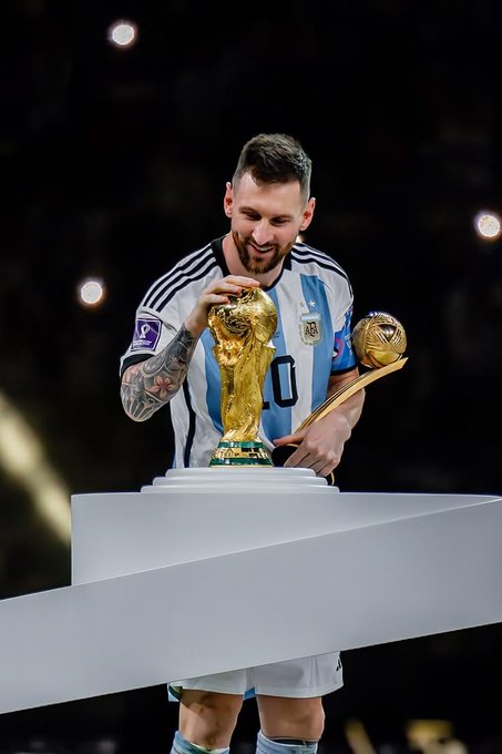 The image features a man wearing a soccer jersey, holding two trophies - one gold and one silver. He appears to be celebrating his achievements by posing for a photo with these awards. The background of the image shows a stadium filled with people, indicating that this is likely an event or gathering related to soccer.