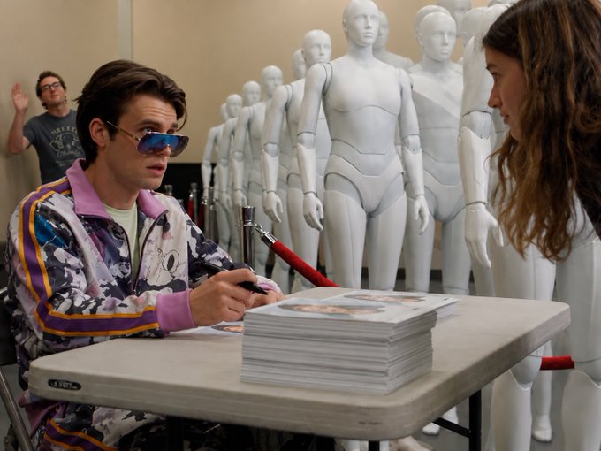 A man wearing colorful patterned clothing and blue sunglasses signs a book or document at a table, with several white mannequins displayed behind him.