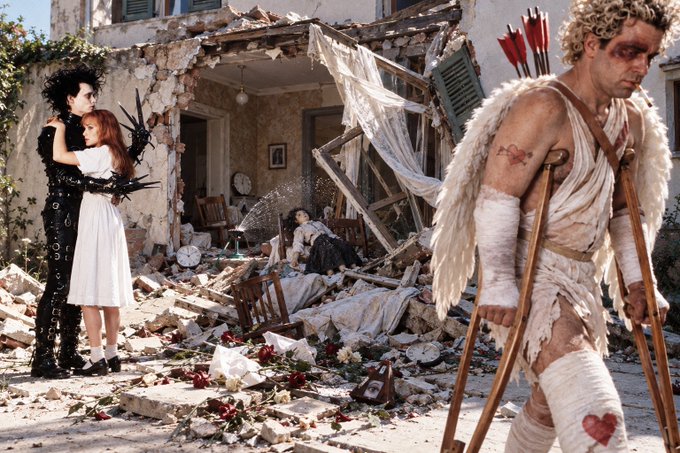 A gothic editorial photo showing a dark-haired figure in black spiked attire embracing a red-haired woman in a white dress, while a wounded angel with bandages and crutches stands in a debris-filled setting with roses scattered about.