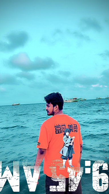 A man with short dark hair wearing a bright orange t-shirt with Japanese text and an anime character graphic, standing by the sea with boats visible in the background under a teal sky.