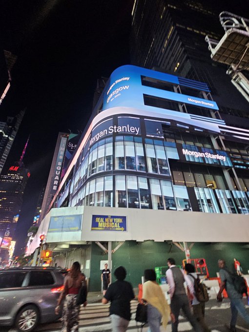 A nighttime view of Times Square in New York City featuring the curved Morgan Stanley building, an H&M sign, and a banner for the 'Real New York Musical'.