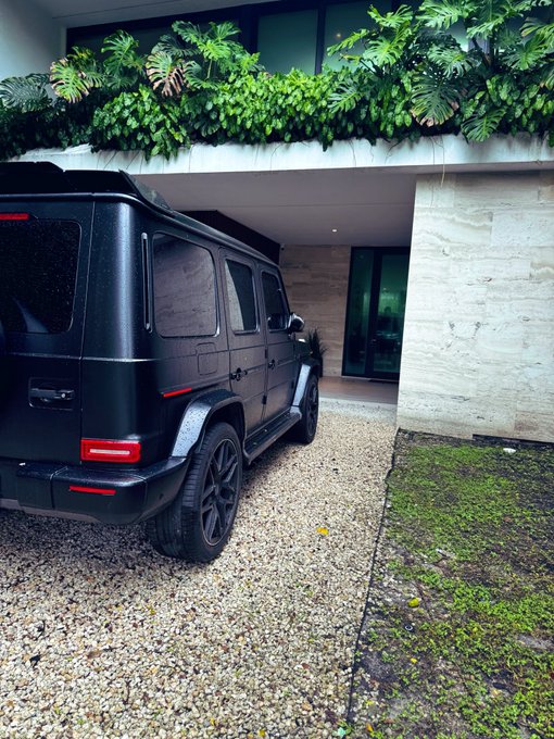 A matte black Mercedes-Benz G-Wagon is parked on a gravel driveway in front of a modern home with lush tropical greenery. The luxury SUV visually embodies the bullish "$AAVE see you at $200" sentiment, projecting wealth and high-end ambition.